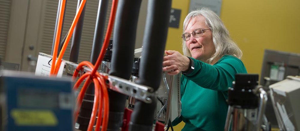 Linda works behind technical equipment and cords at ϲʿͼ.