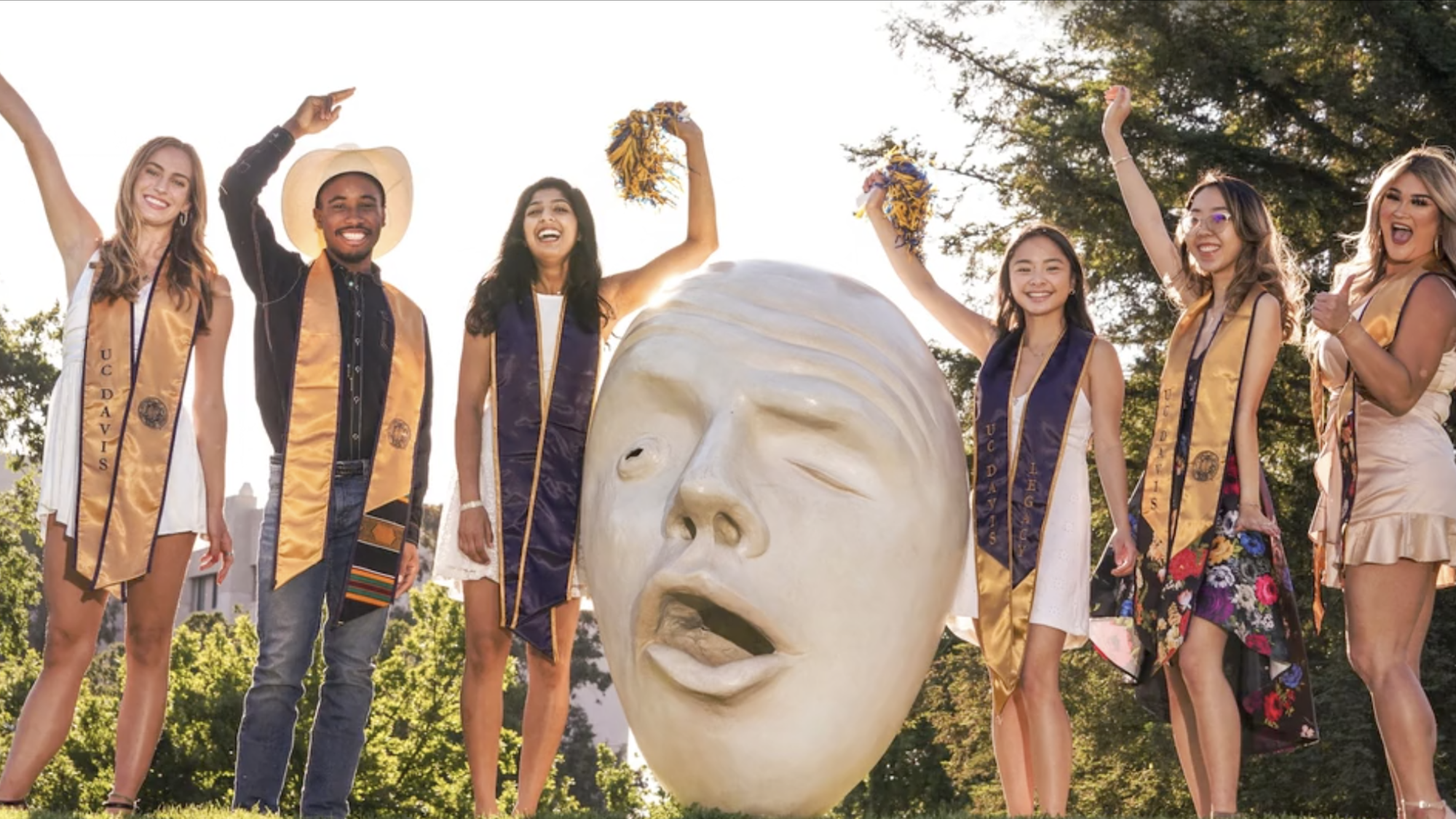 six students in graduation hats and sashes pose near an Egghead statue at ϲʿͼ