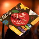 A grad cap that reads, "And the student became the teacher," in white cursive within a bright red apple, surrounded by teaching equipment like rulers, crayons and a pencil that reads, "Ms. Murillo." The photo was taken at ϲʿͼ' School of Education Commencement graduation ceremony in 2023.