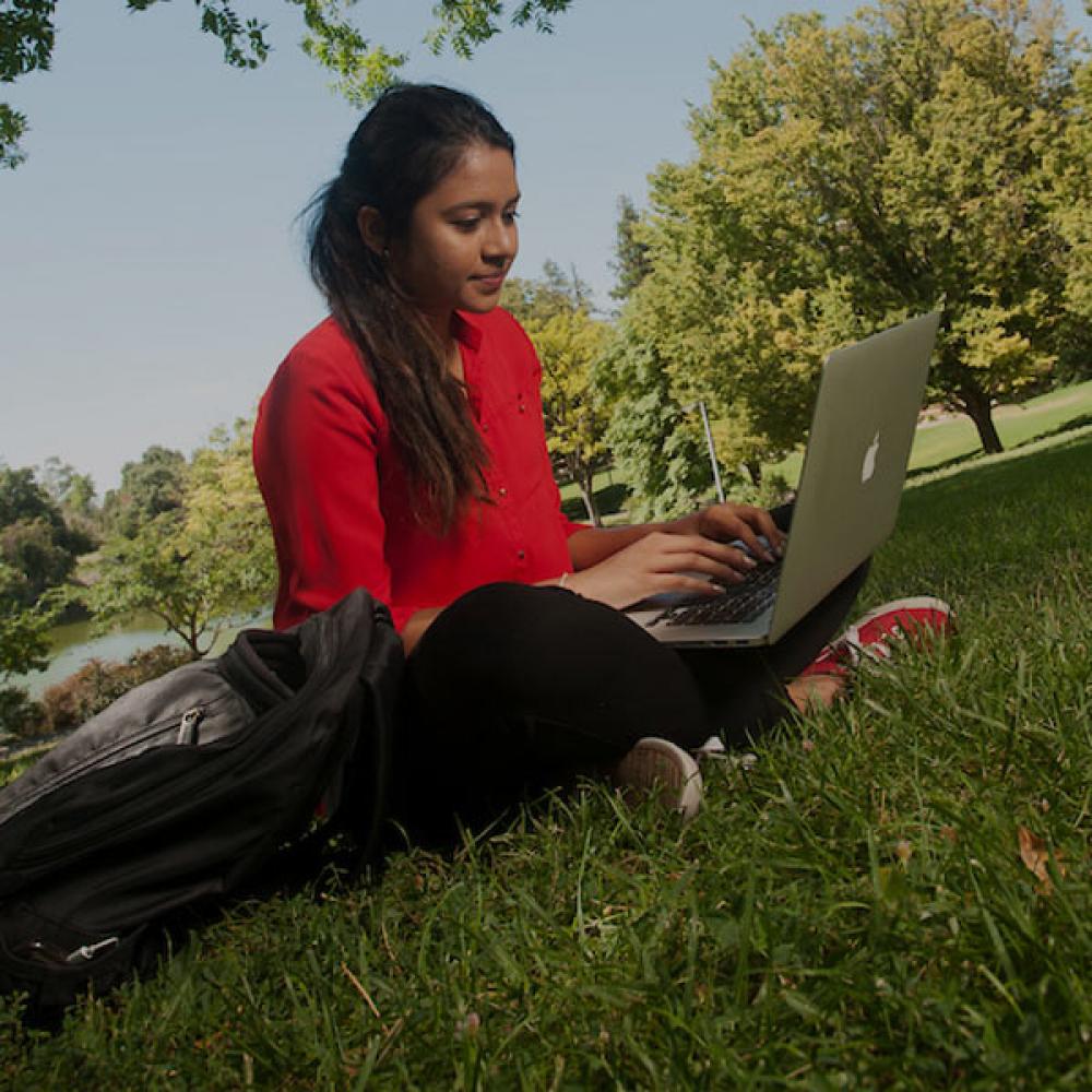 A female student in red reads from her laptop on the ϲʿͼ quad