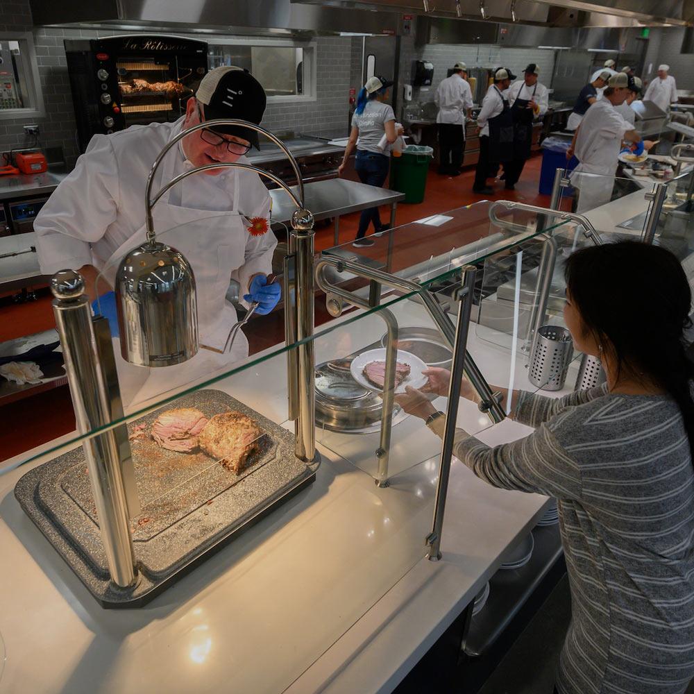 A dining commons employee serves a cut of meat to a ϲʿͼ student