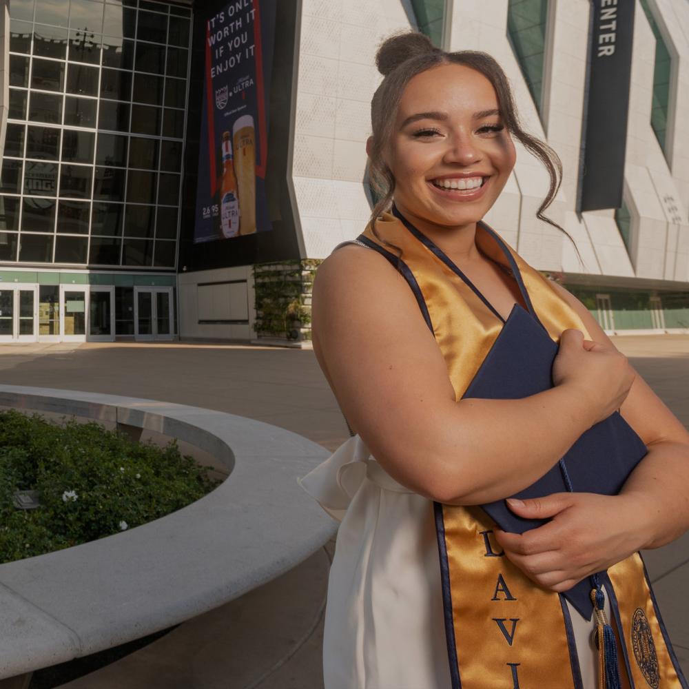 A ϲʿͼ student standing outside of the Golden 1 center holdint their grad regalia